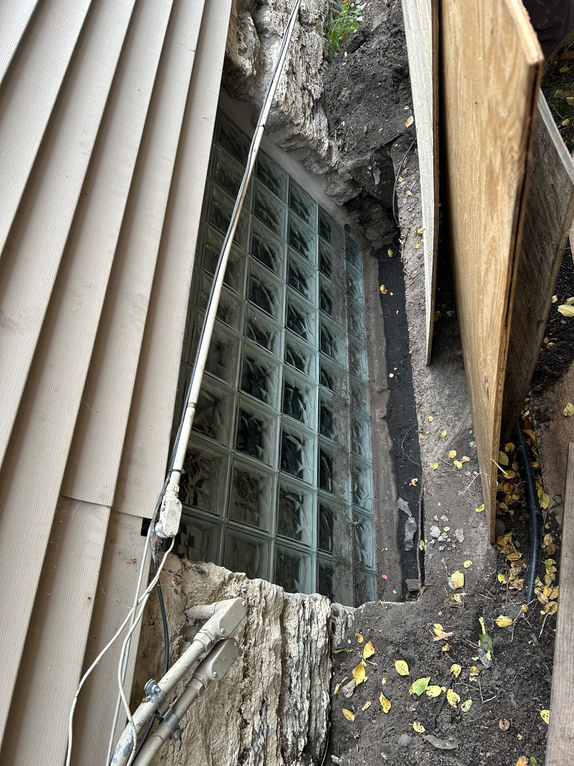 Glass block window in a narrow trench between siding and a wooden wall. Earth and debris surround it.