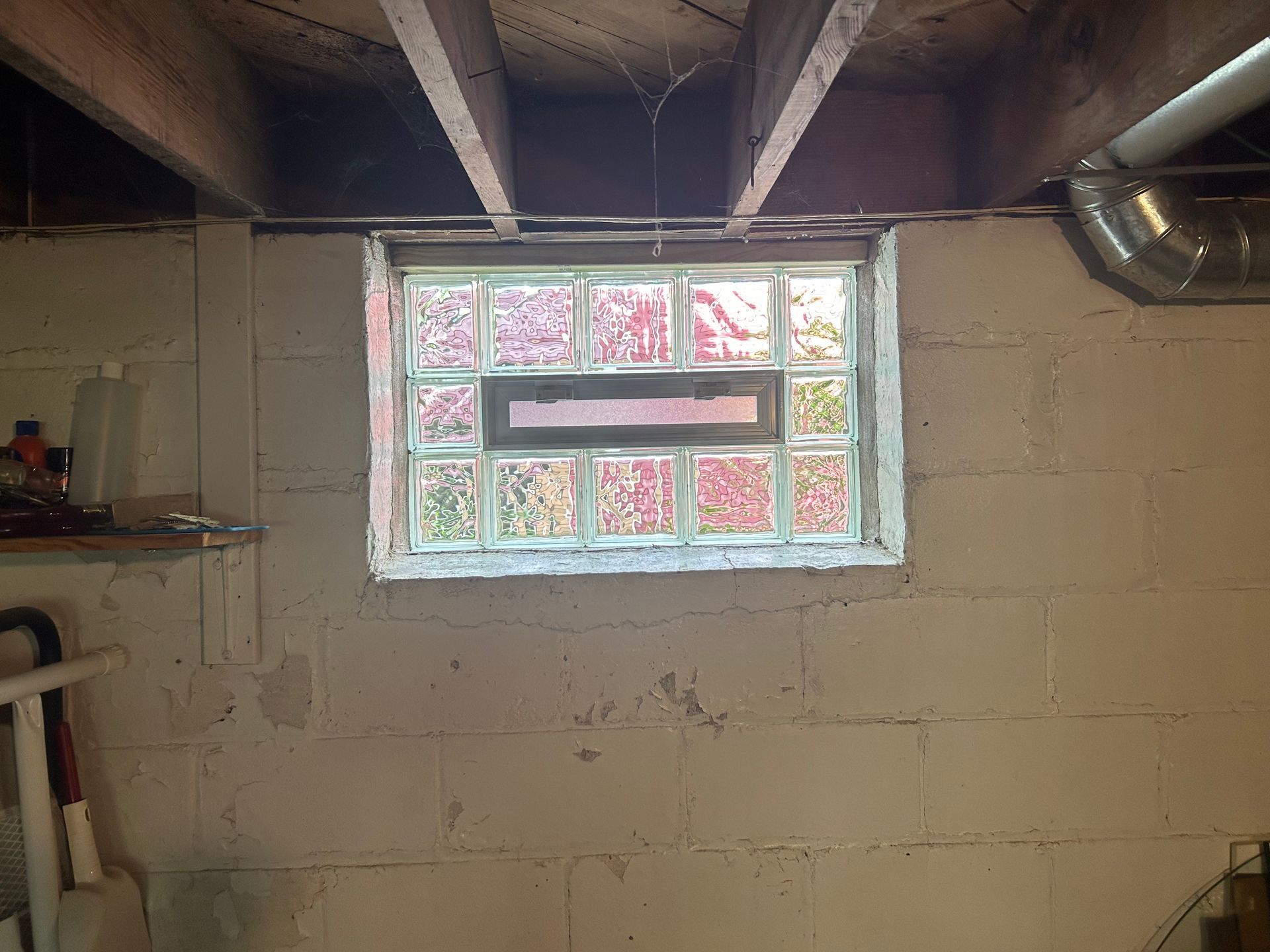 Basement wall with glass block window. Gray cinder blocks, white window frame, exposed beams, ductwork.