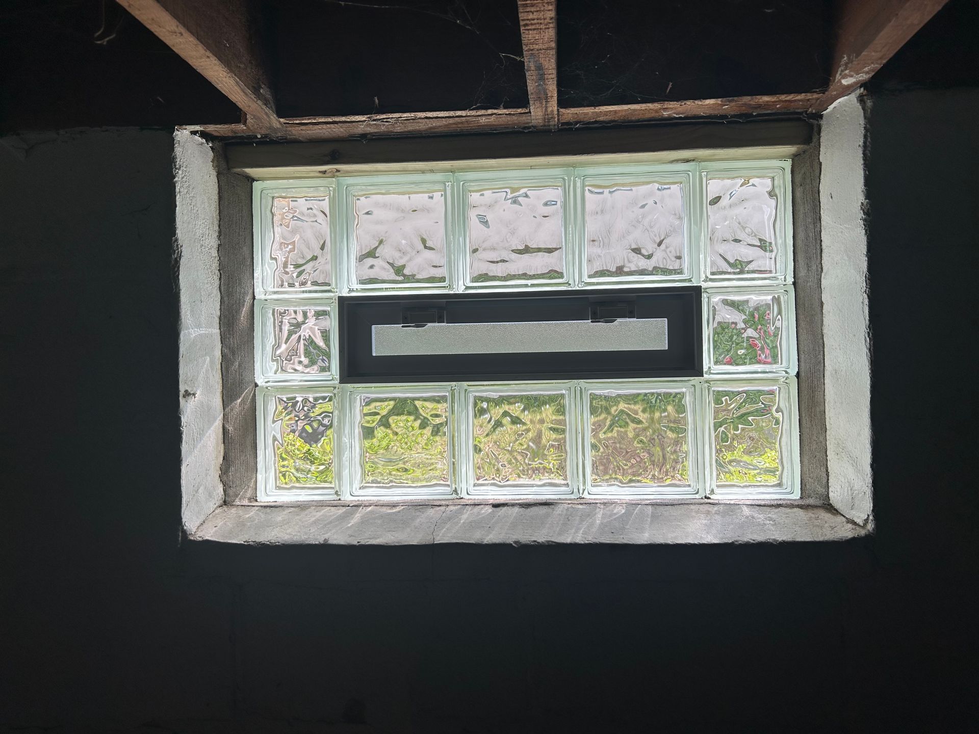Glass block window in a dark, concrete wall. Light shines through, with some foliage visible.