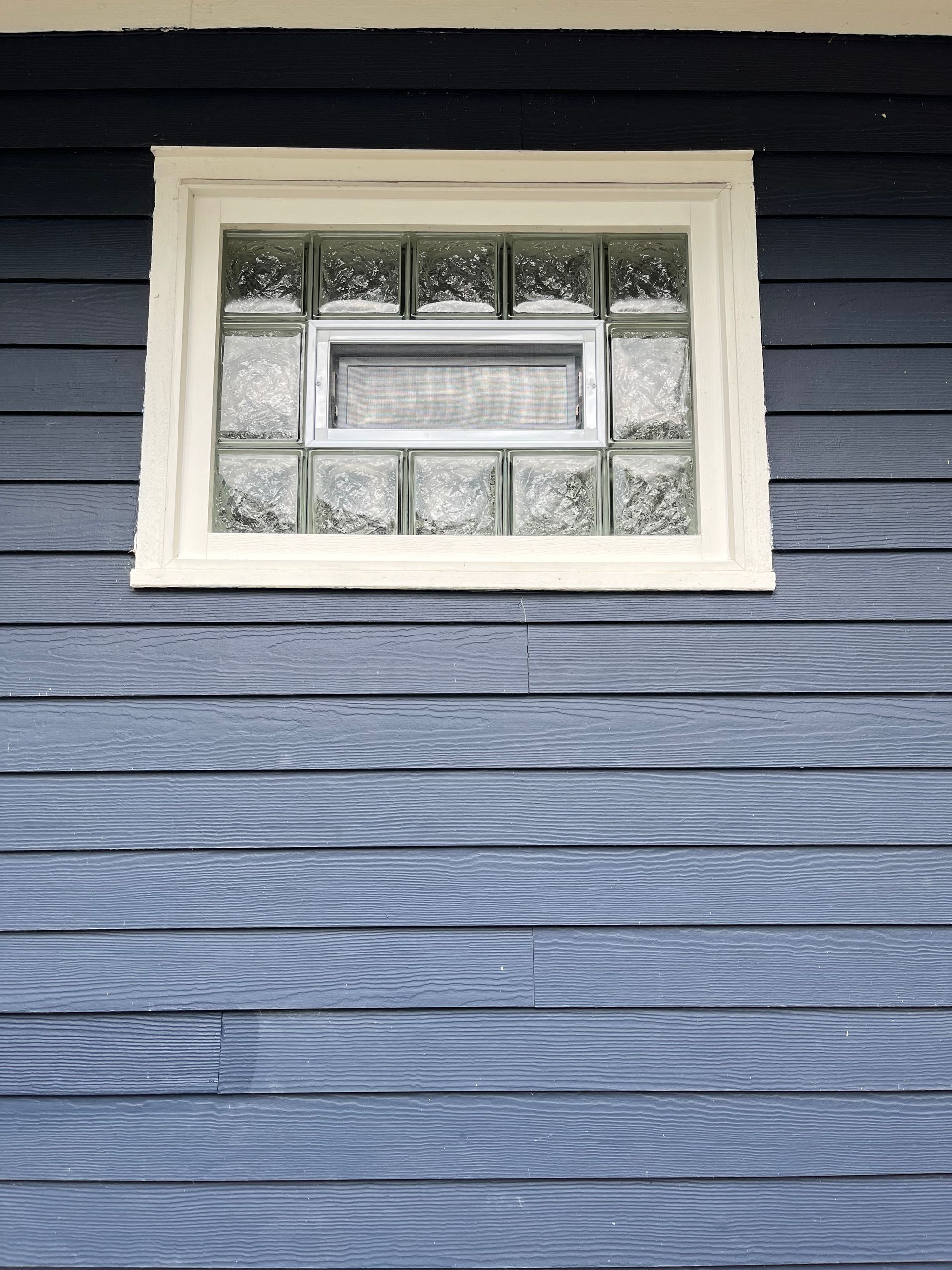Glass block window with a small opening, set in a white frame on a blue-painted wood exterior.