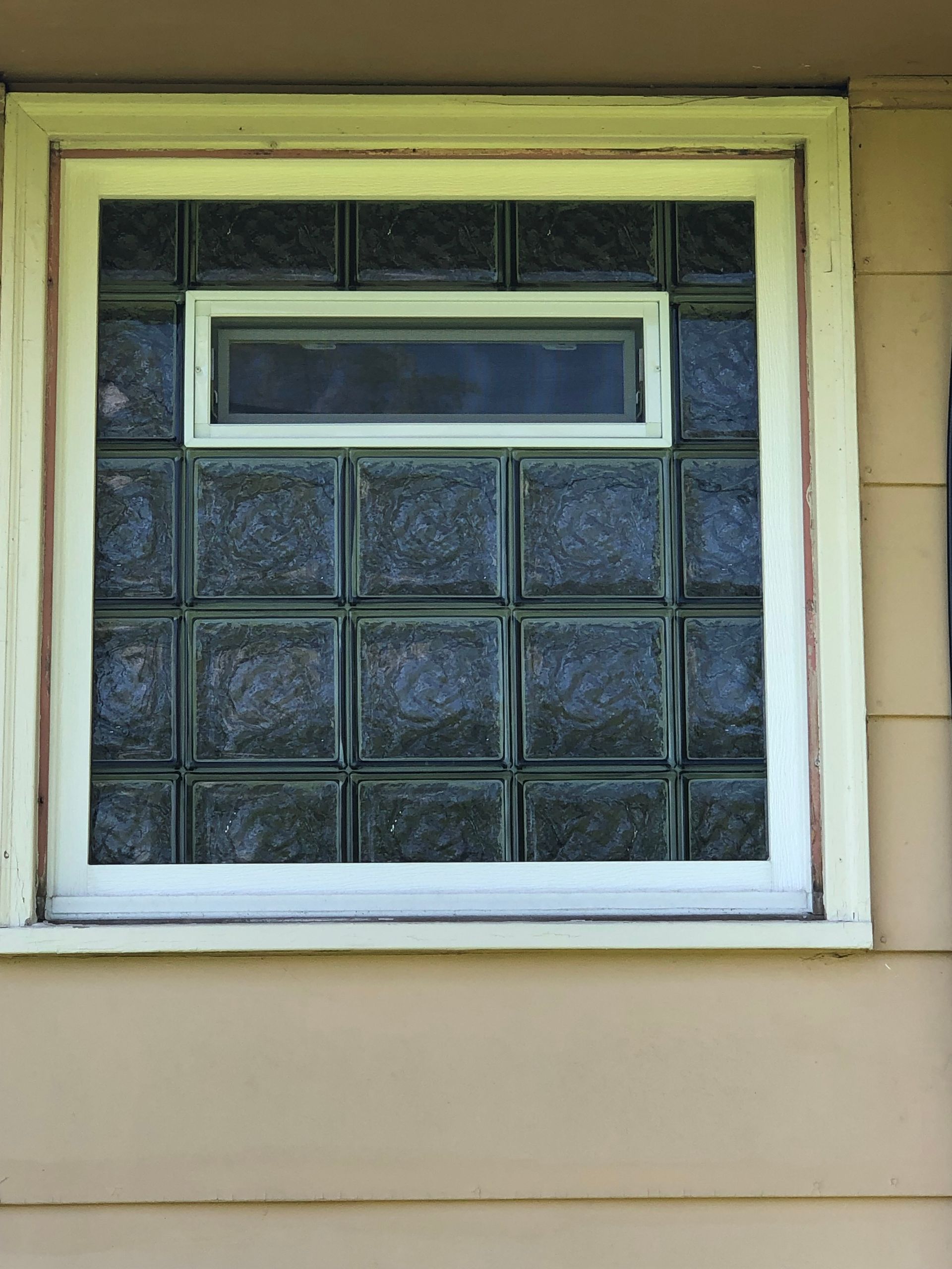 Glass block window with small rectangular window centered in the middle, surrounded by a white frame on a tan wall.