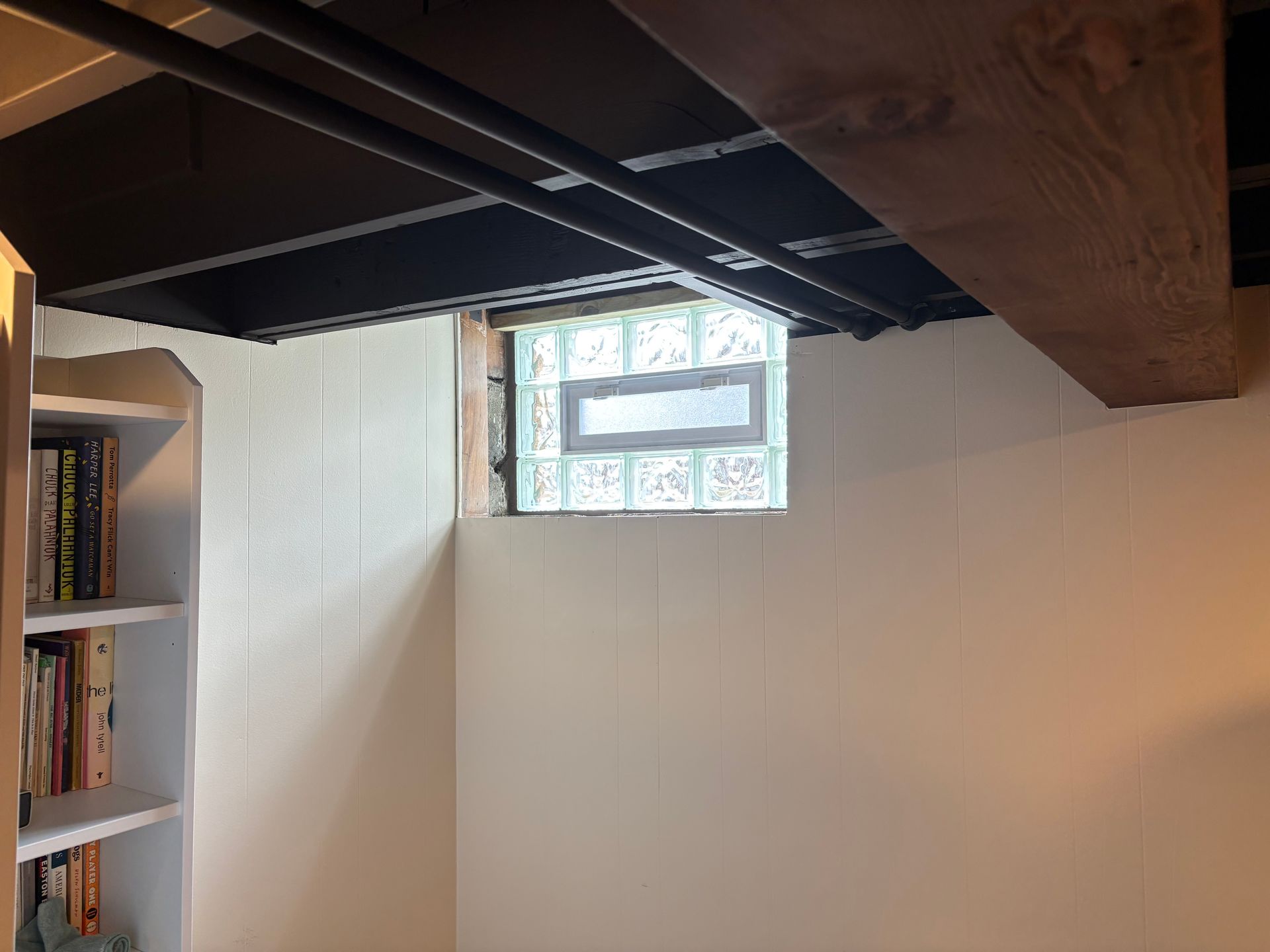 Basement interior with a glass block window, white paneled walls, and exposed ceiling beams. A bookshelf is in the corner.