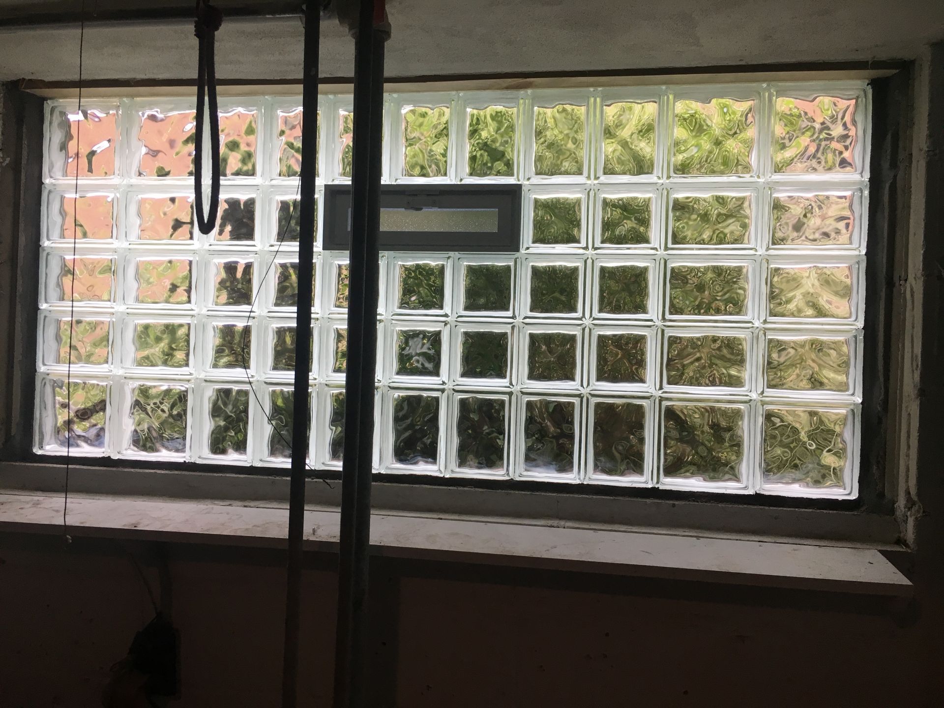 Glass block window, interior view. Blocks are translucent; greenery visible through them. Grey frame.