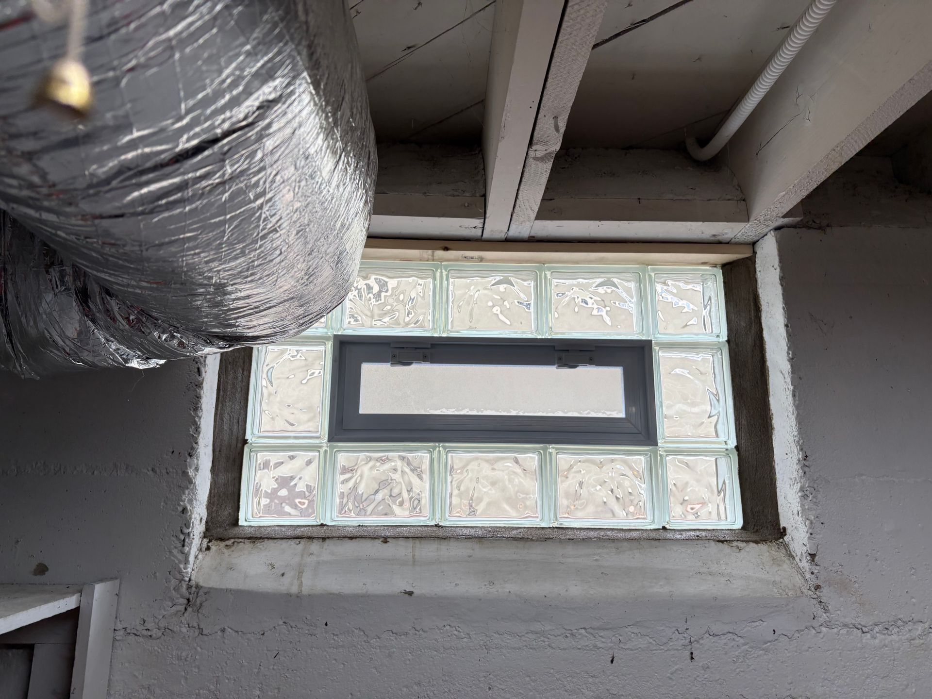Glass block basement window with a rectangular vent, set in a concrete wall, and ductwork above.
