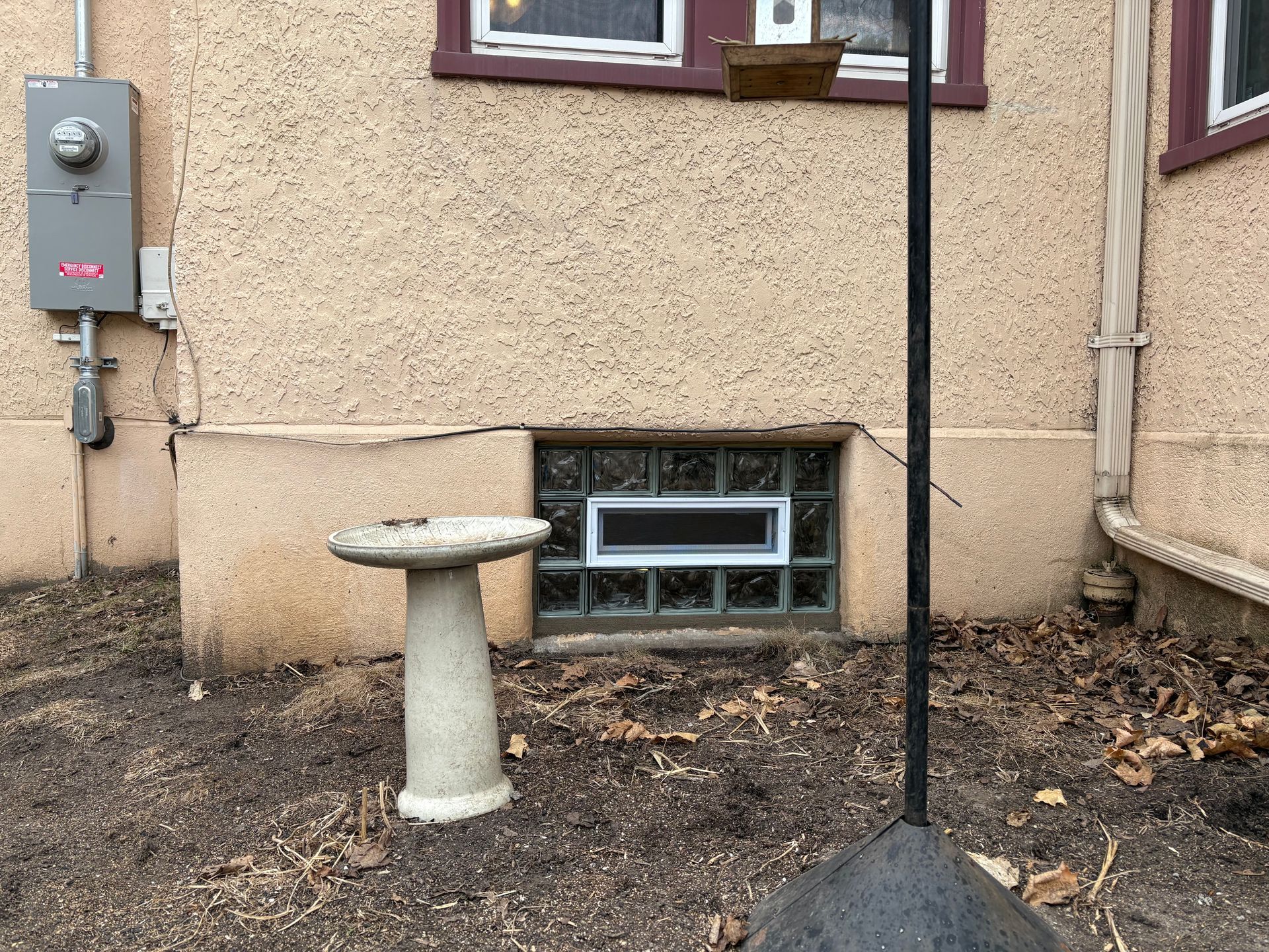 Birdbath near a beige stucco house with a window and electrical box; dry yard.
