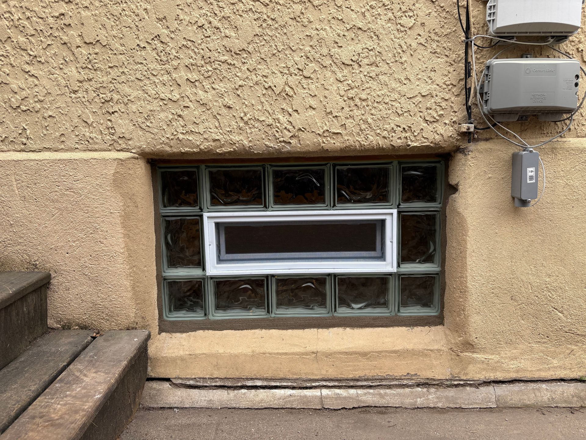 Window with glass blocks in a beige stucco wall; basement steps on the left; electrical boxes on the right.