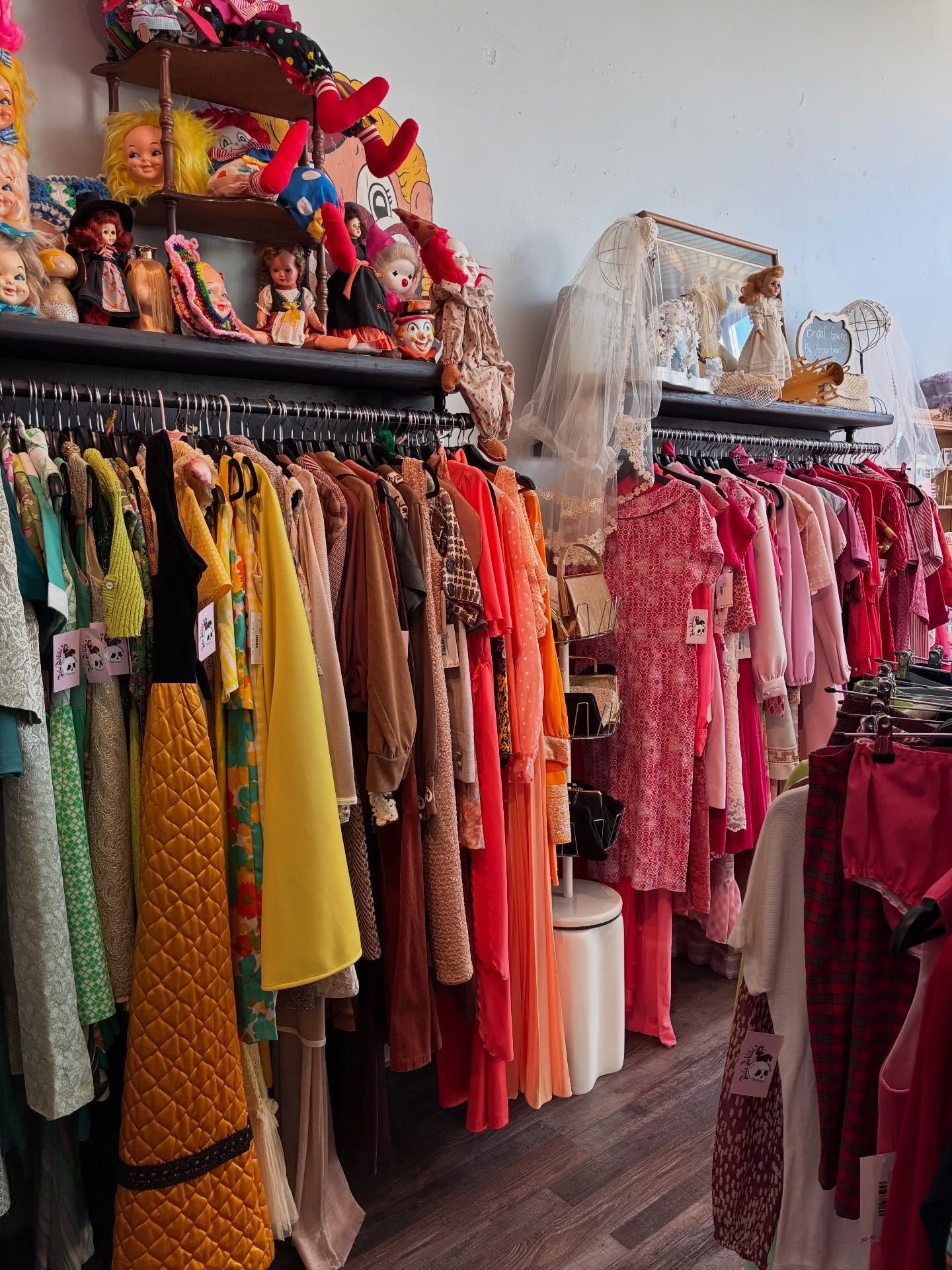 Clothing racks in a vintage clothing store, with shelves of dolls and decor overhead
