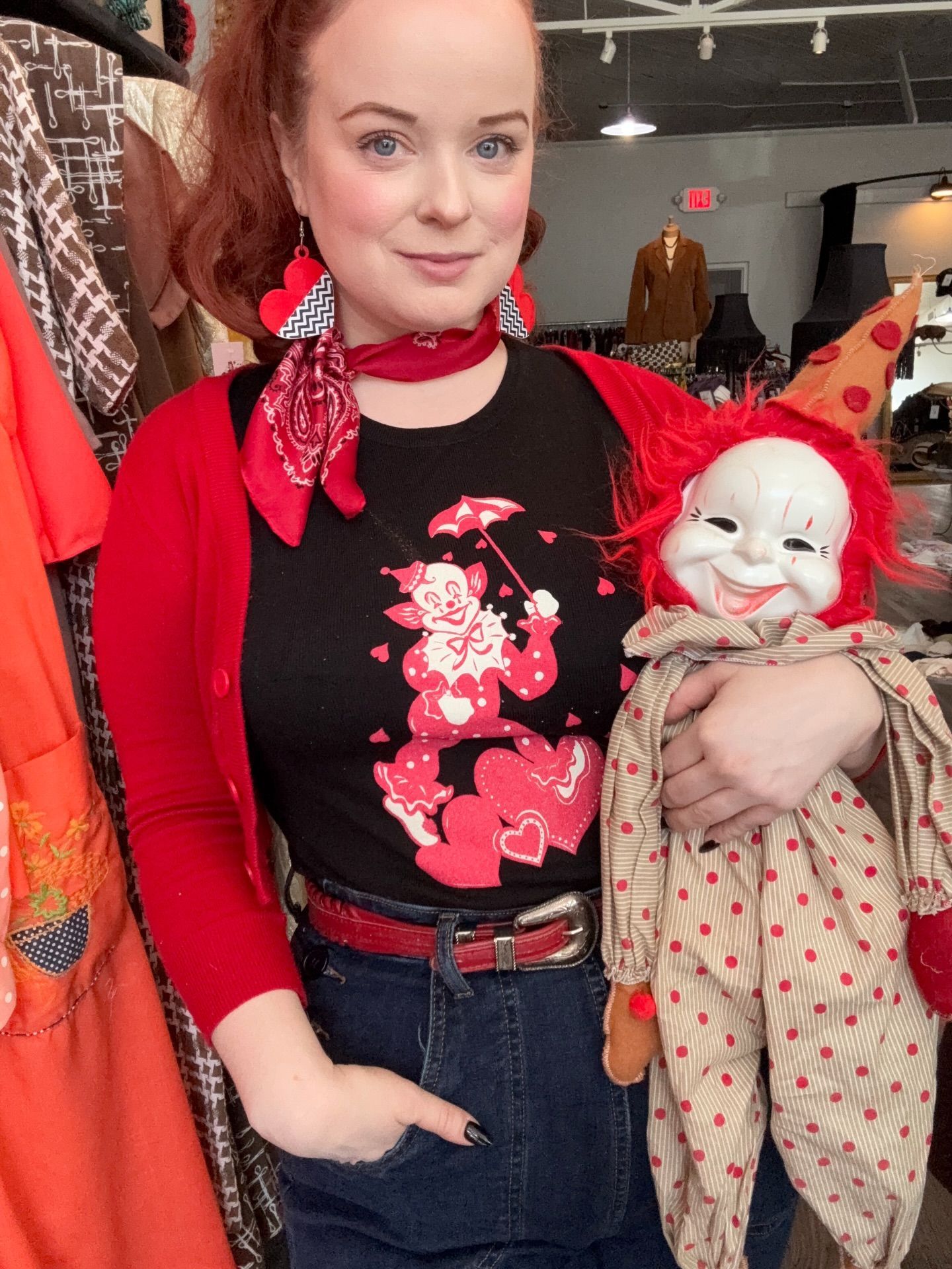 Woman in red, black top with clown graphic, holding clown doll in a vintage shop