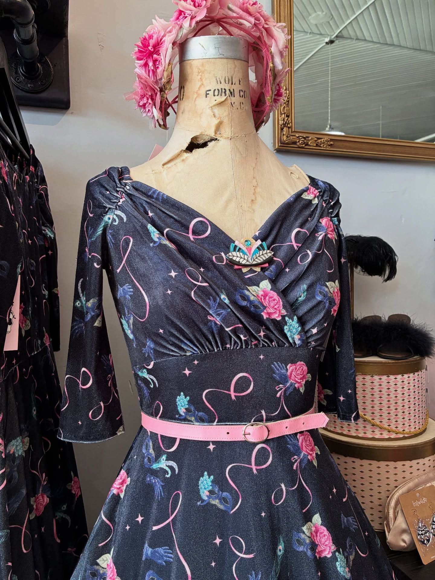 Mannequin in vintage floral dress, pink belt, and feathered hat, displayed in a shop setting