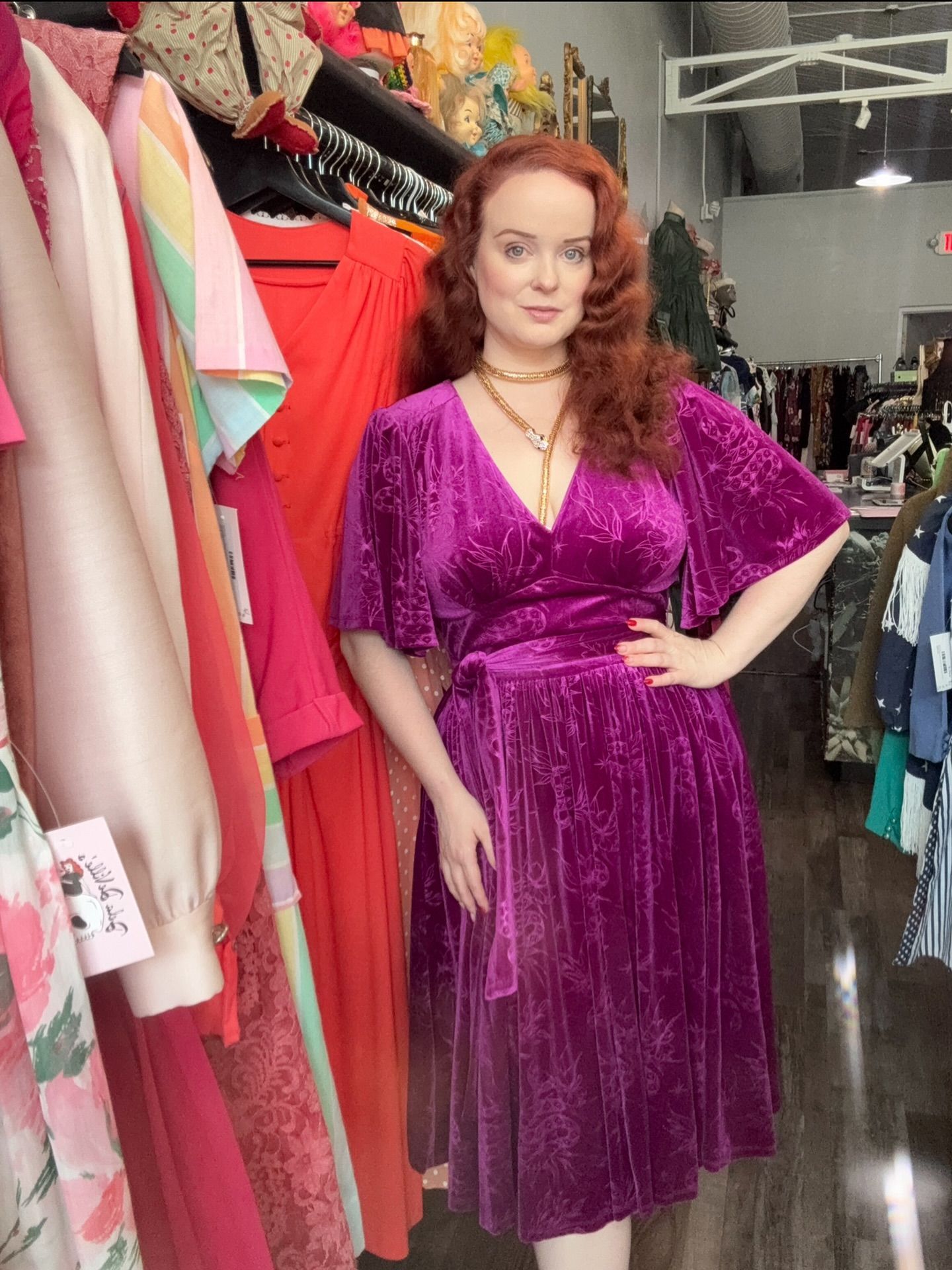 Woman in a purple velvet dress in a vintage clothing store, posing with a hand on hip