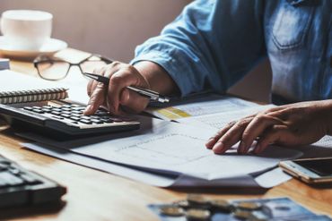 A person working on finances with a calculator, paperwork, and a pen at a desk.