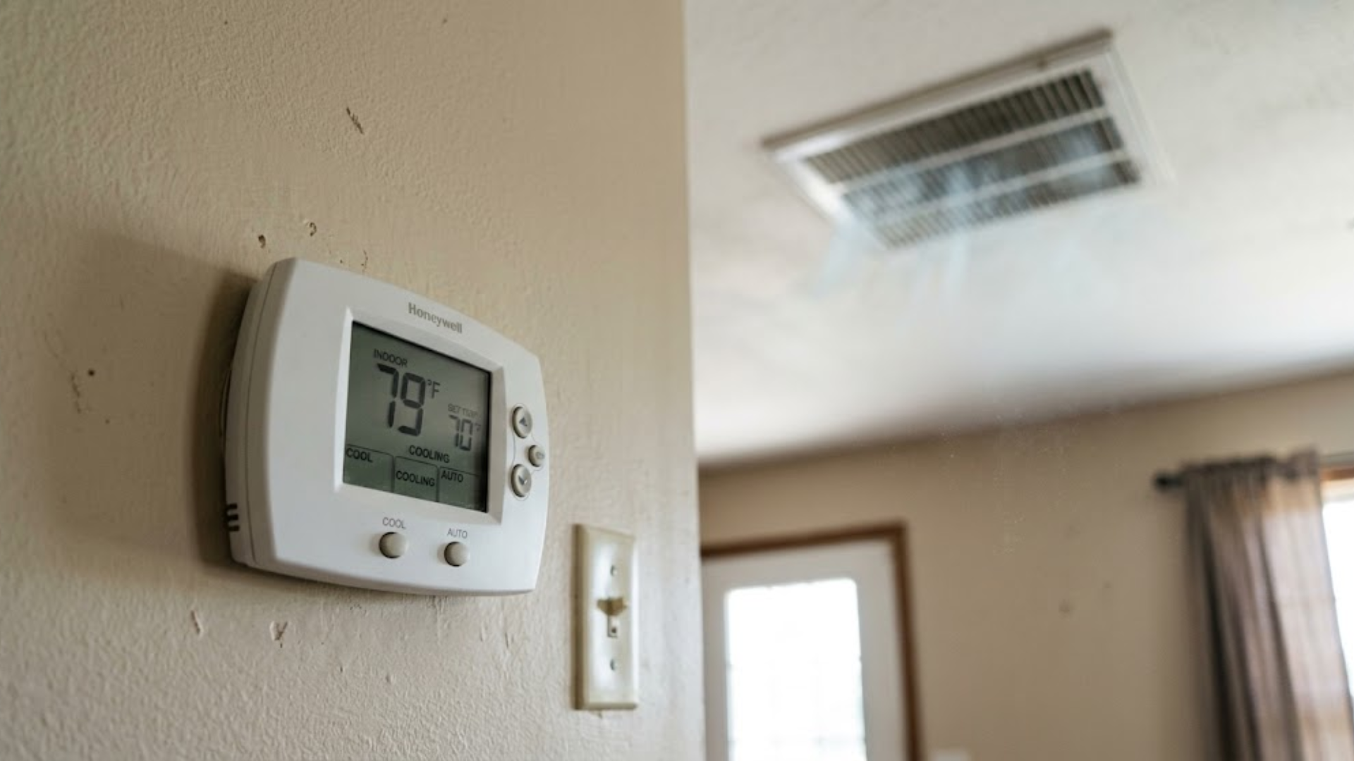 Wall-mounted thermostat on a beige wall near a light switch, with a ceiling vent and doorway in the background.