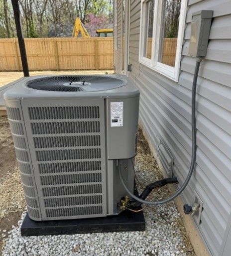 Outdoor air conditioner unit beside a house on gravel, with a fenced yard in the background.