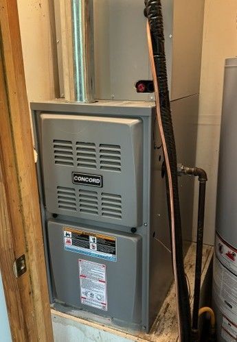 Gray furnace unit in a narrow utility closet beside exposed pipes and ductwork.
