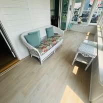 Sunroom with a white wicker sofa, matching side table, and light-colored flooring. Sunlight streams through windows.