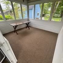 Empty sunroom with brown carpet, white paneled walls, and a wooden table near windows.