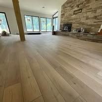 Empty room with light wood flooring, a fireplace, and large windows. Sunlight streams in.