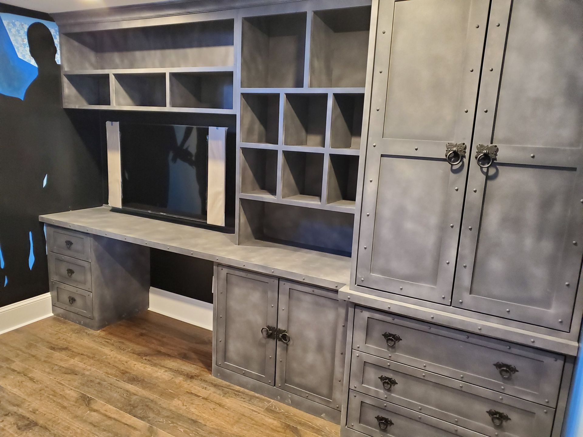 Built-in gray office shelving with desk, cabinets, and storage, against a black wall and wood floor.