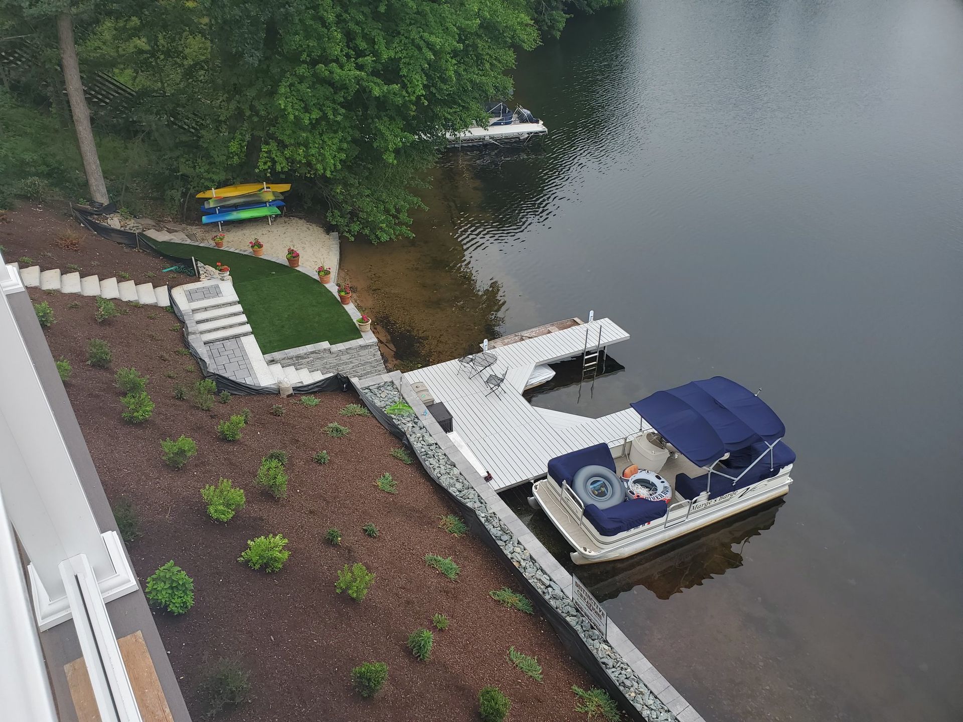 A pontoon boat docked at a white pier on a lake. Landscaped shoreline with steps and trees.