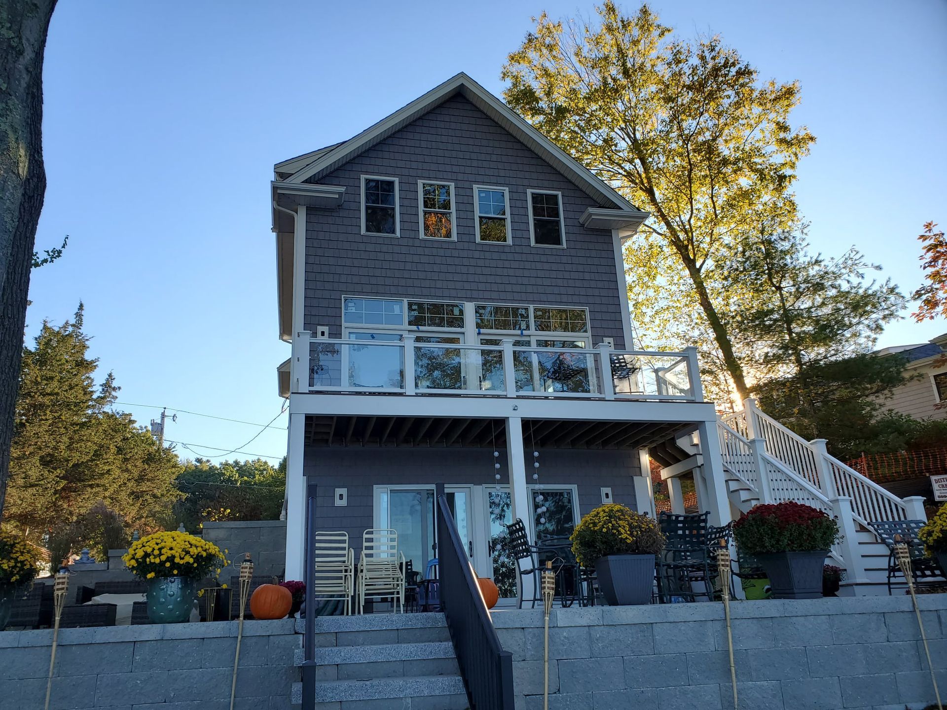 Three-story gray house with balcony overlooking water, stairs, potted mums, and a tree.