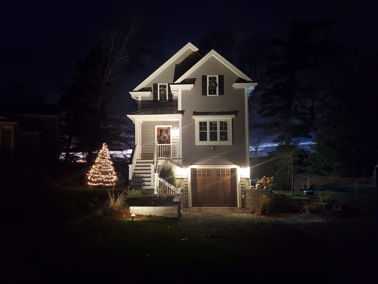 House at night with Christmas lights. Gray siding, lit Christmas tree.