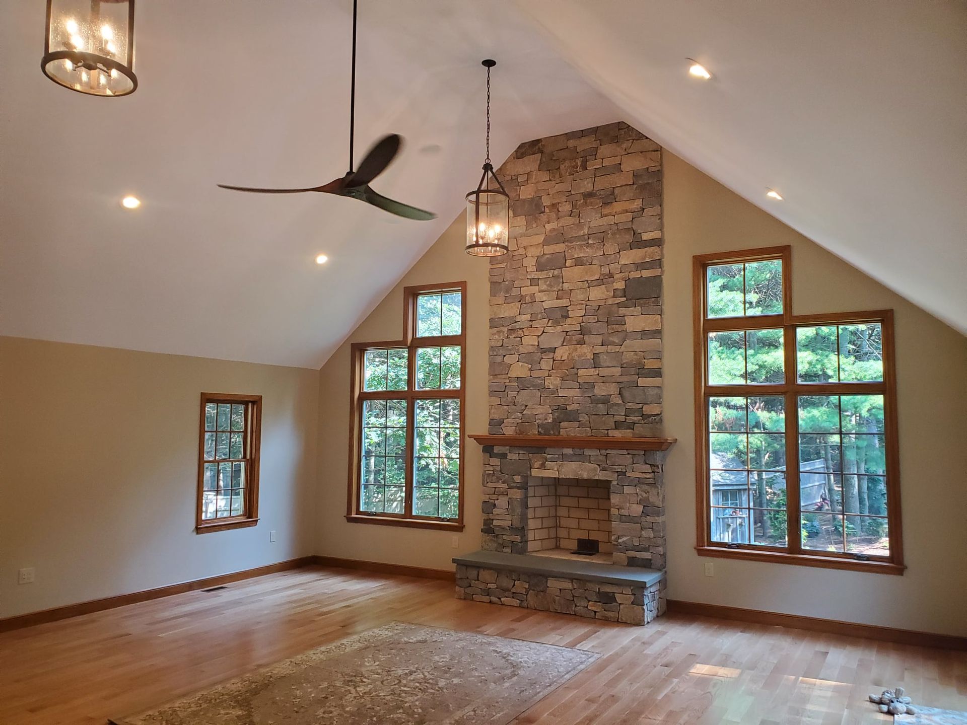 Spacious living room with stone fireplace, large windows, wood floors, and a high ceiling.