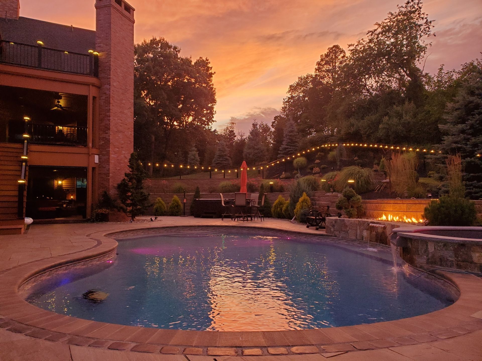 Pool and outdoor living space at sunset: pool, fire pit, house with balcony, string lights, colorful sky.