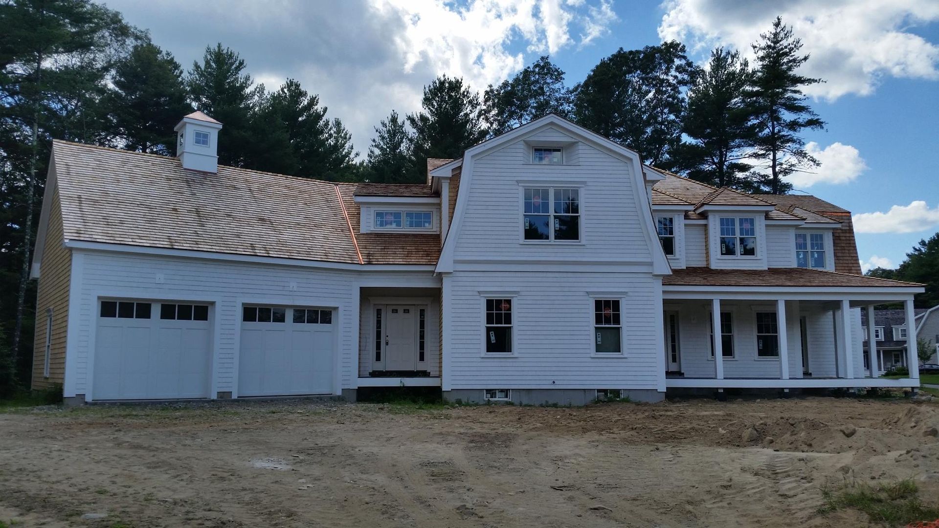 White house under construction with a porch, garage, and cedar shake roof; set in front of trees.