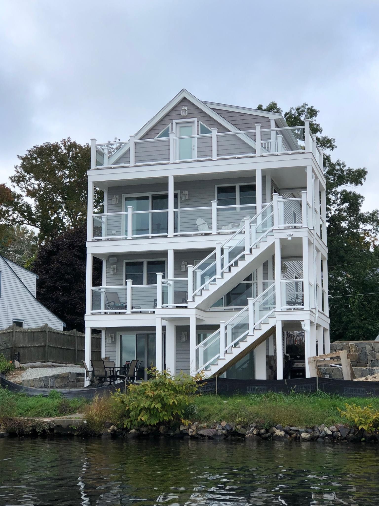 Three-story gray and white house with glass-railed balconies and outdoor stairs, overlooking water.