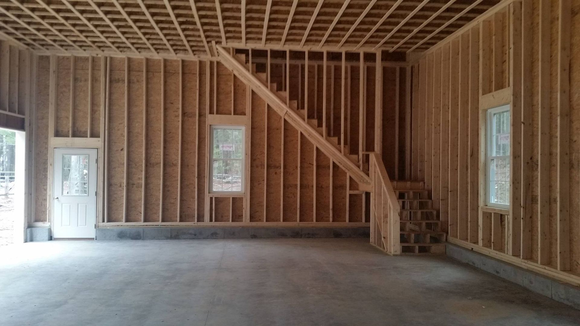 Interior view of a building under construction; wood framing, staircase, concrete floor, windows, and door.