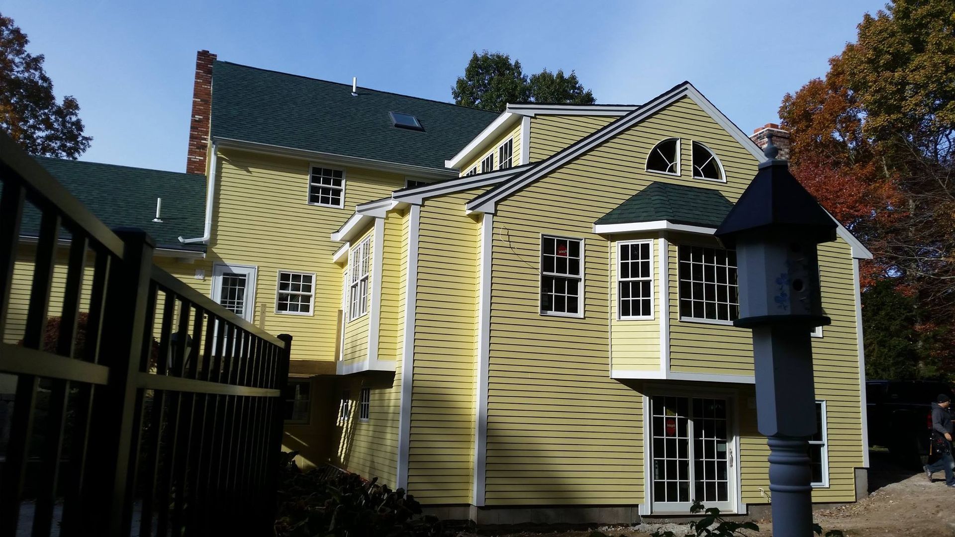 Yellow house with green roof, multiple windows, and a birdhouse, sunny day.