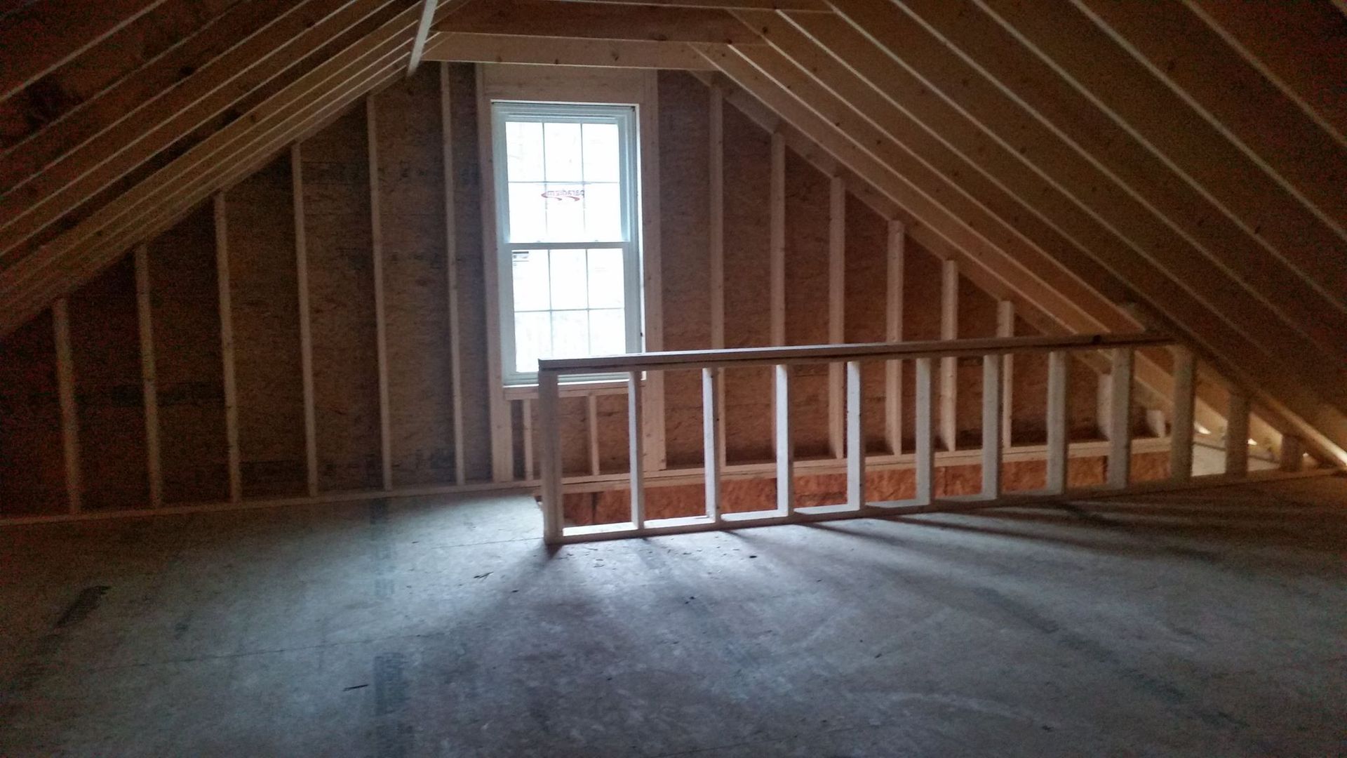 Unfinished attic with sloped walls, railing, window, and floor. Sunlight streams through the window.