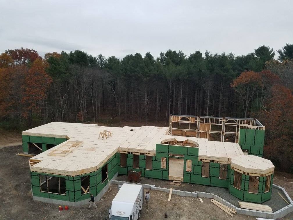 House under construction in a forest setting, with wooden frame and green sheathing visible.
