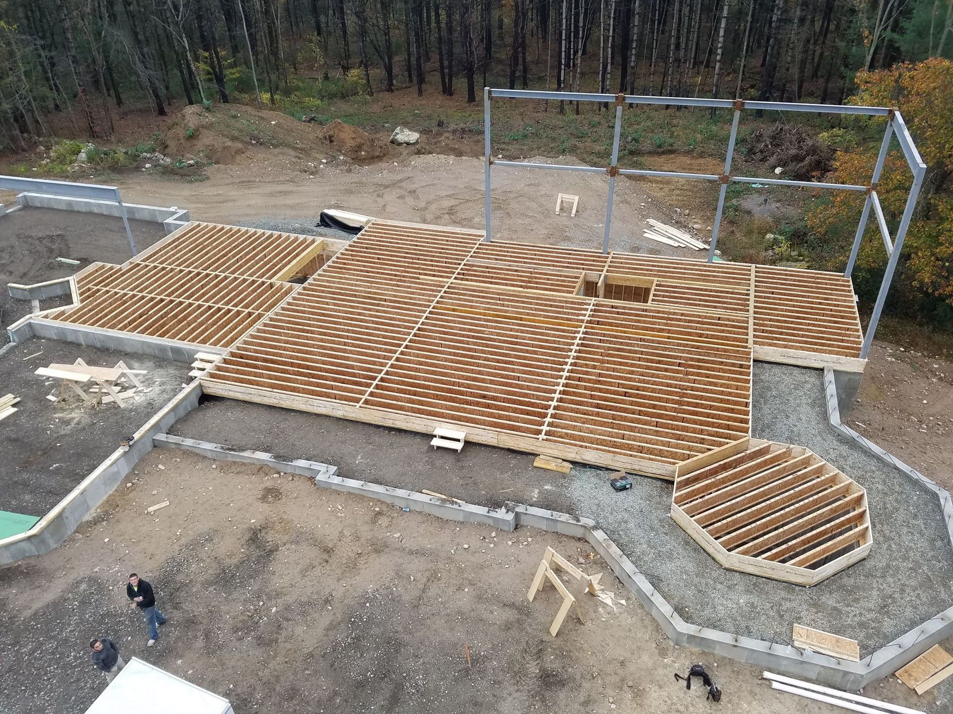 Aerial view of a wooden deck under construction on a gravel foundation; workers nearby, metal framing in background.