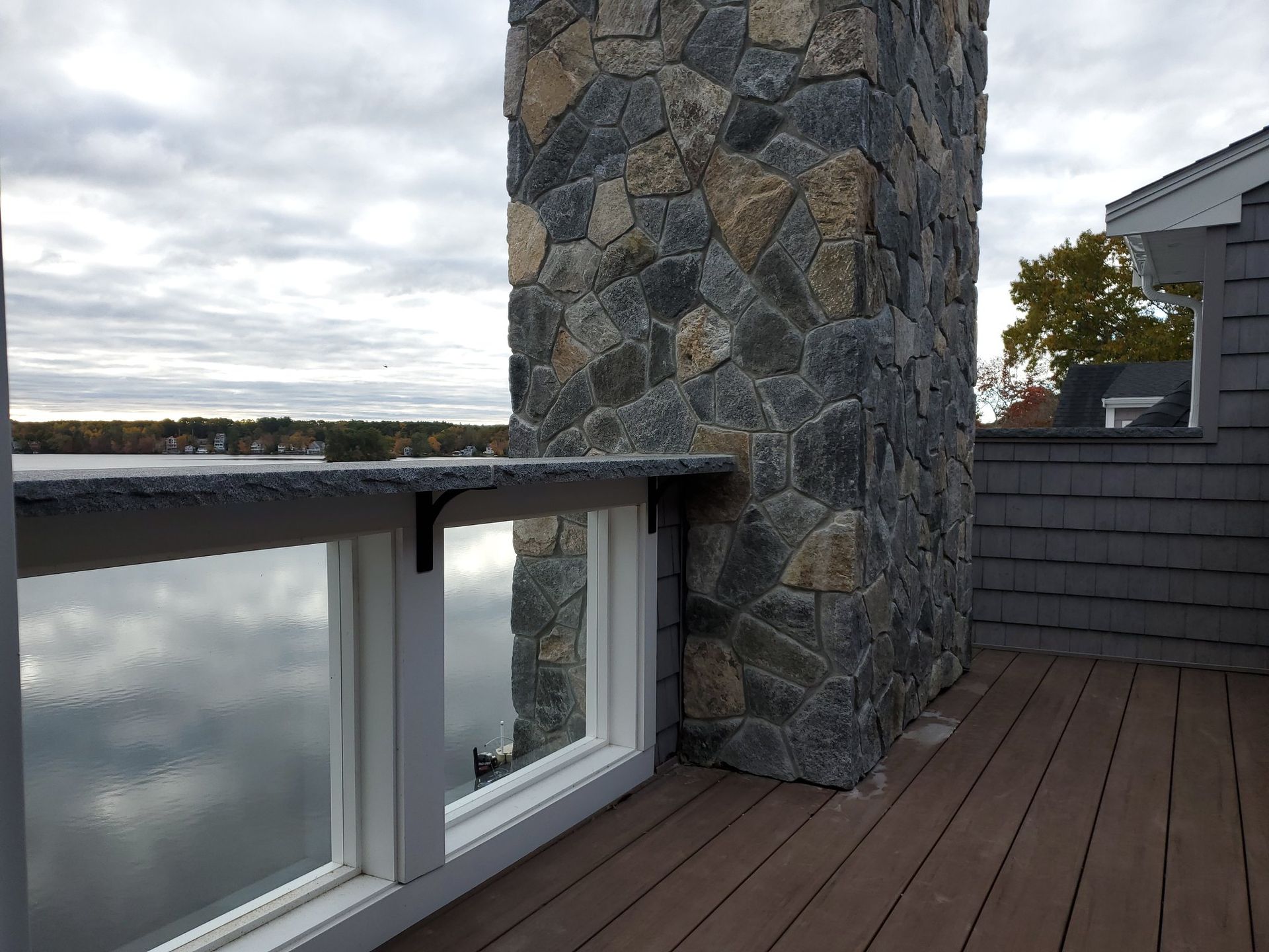 Stone chimney on a deck with a lakeside view. Gray and brown stone, with windows and composite decking.