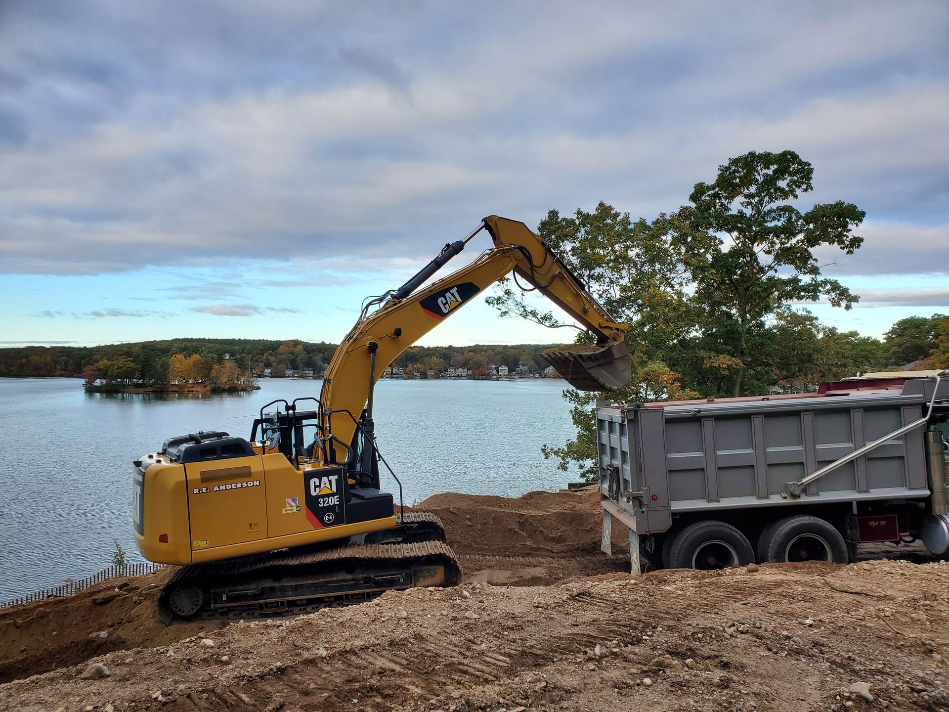 Yellow excavator loading dirt into a gray dump truck near a lake.