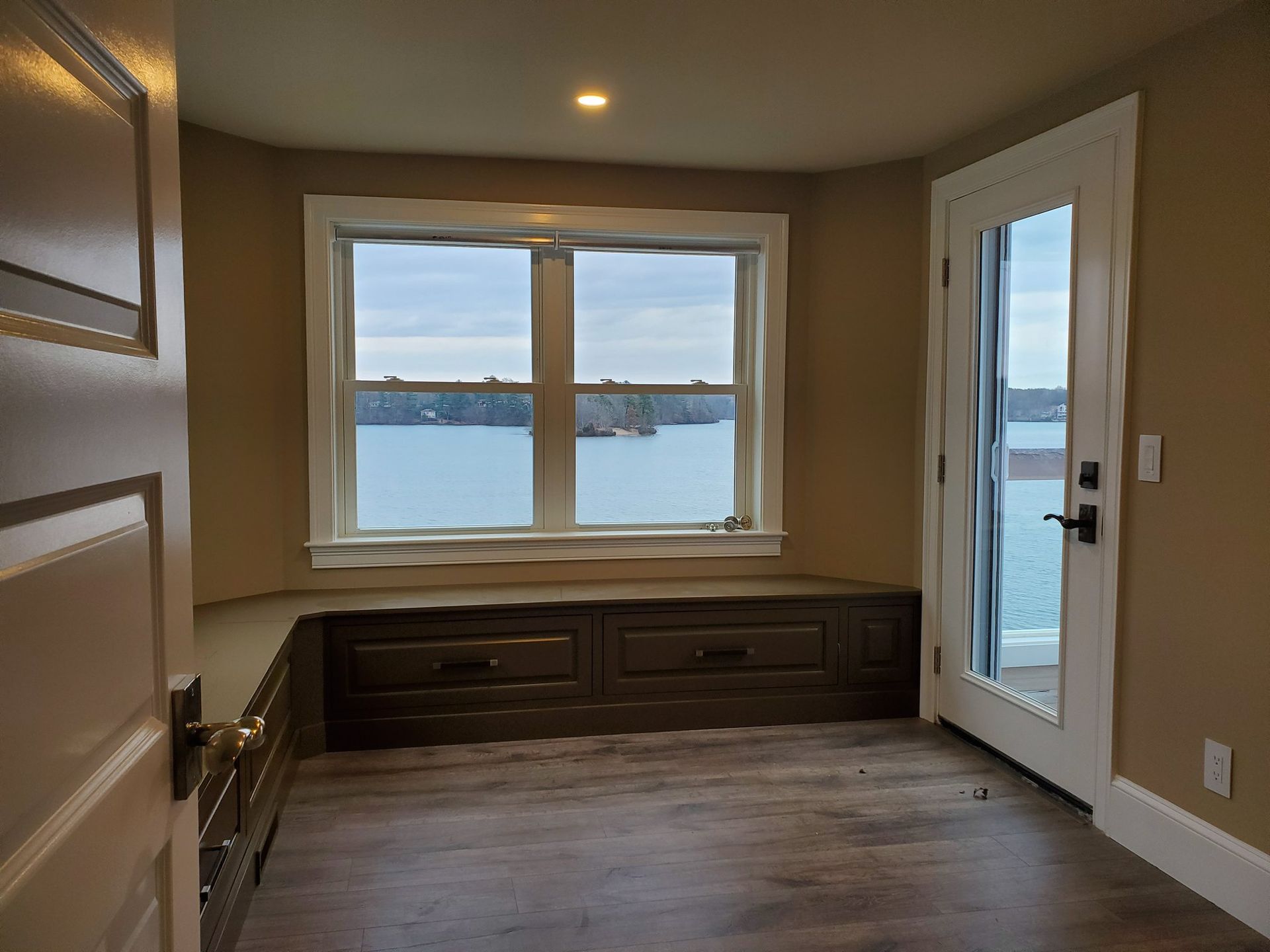 Cozy room with window seat and water view; a door leads to a balcony, brown cabinetry, and beige walls.