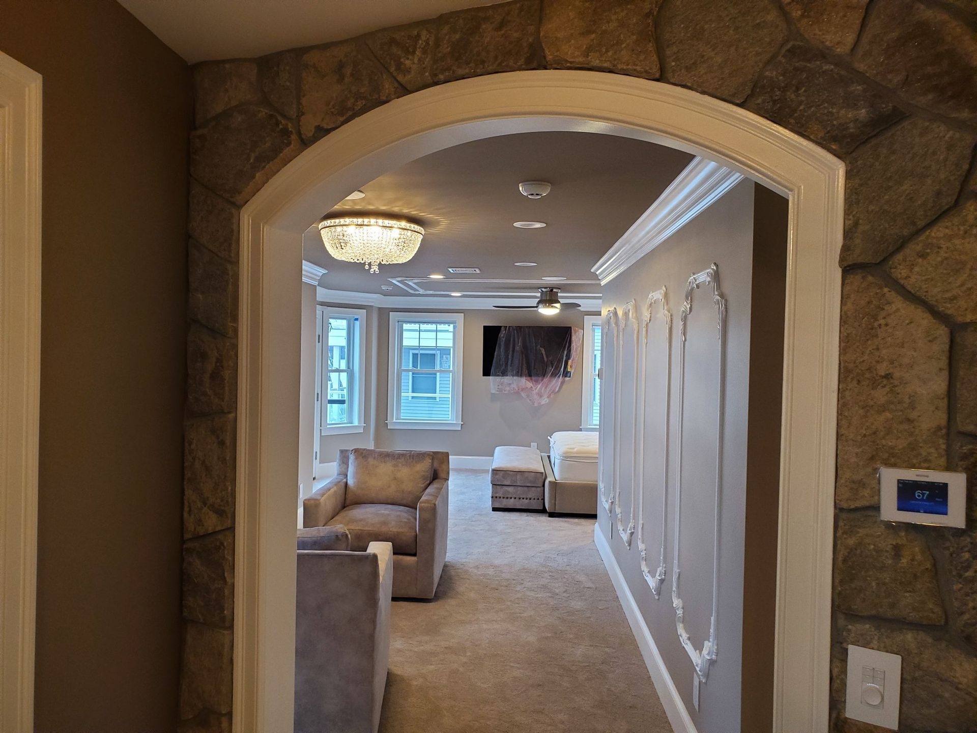An arched doorway with stone facing leads to a bedroom with armchairs, art, and a chandelier.