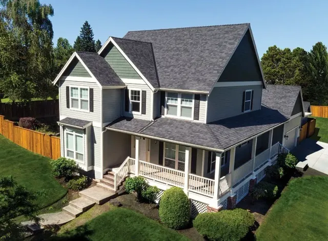 Gray two-story house with a wraparound porch and dark roof in a sunny suburban yard