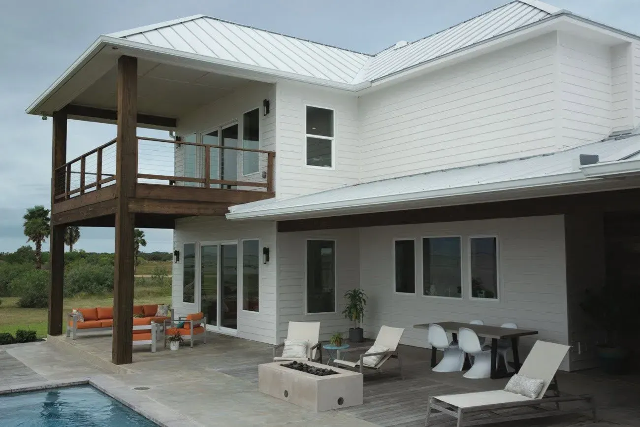 Modern white two-story house with a balcony, patio lounge chairs, and a poolside deck