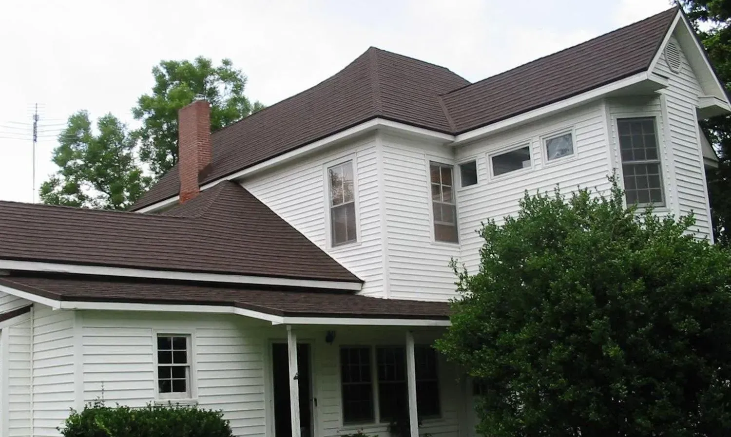 White two-story house with brown shingle roof and a front porch surrounded by trees