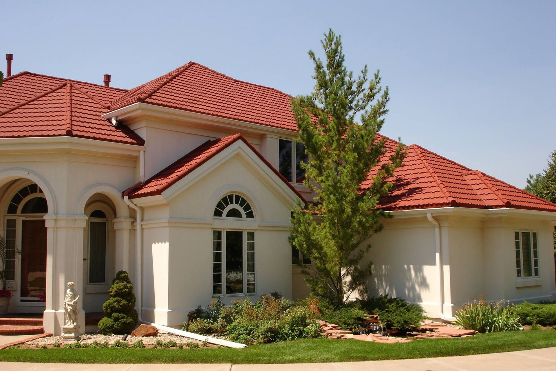 Large cream-colored house with a red tiled roof and arched windows, fronted by a manicured lawn and shrubs