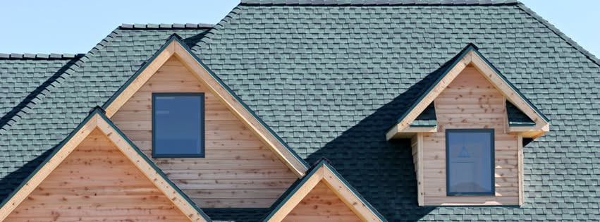 Close-up of a house roof with gray shingles, wooden trim, and two dormer windows against a blue sky