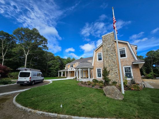 Stone house with U.S. flag, white van, and green lawn under a blue sky