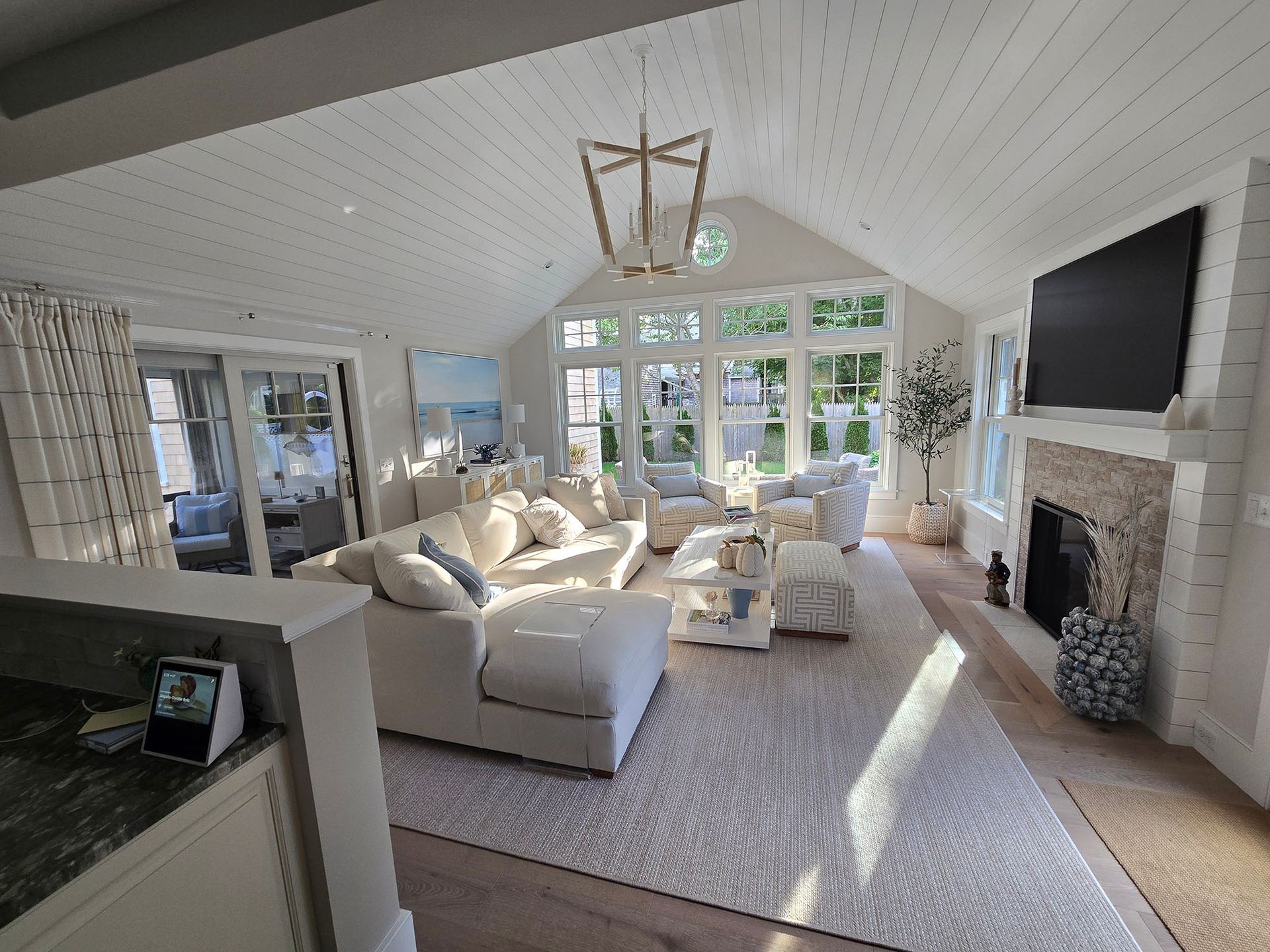 Bright living room with white sofas, stone fireplace, large windows, and a TV on the wall.