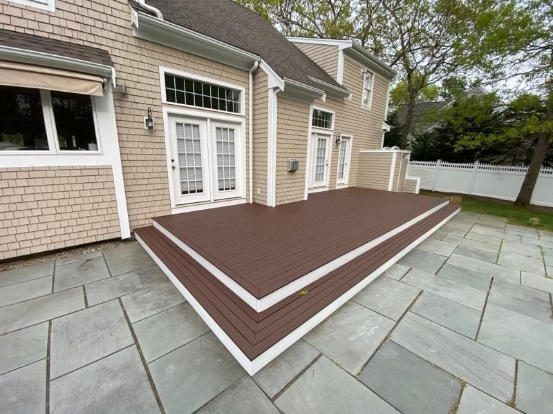 Backyard deck with brown wood surface, white trim, and stone patio outside a house
