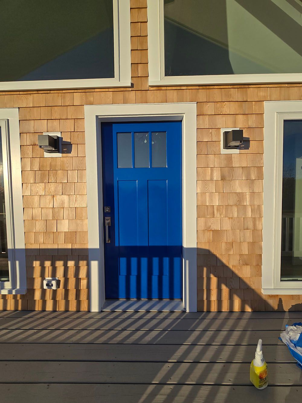Blue front door on a shingled house with windows, striped shadows, and a small bottle on the porch.
