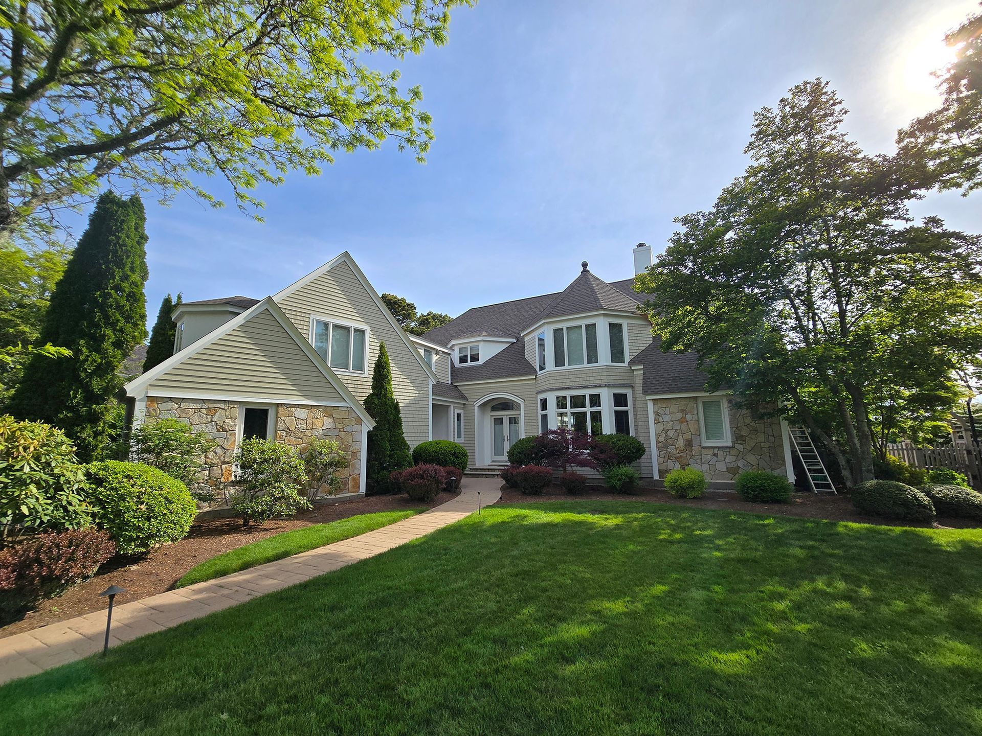 Large suburban stone house with gabled roof, front lawn, trees, and a sunny sky