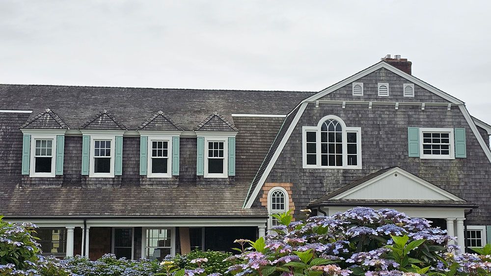Gray shingled house with white-trim windows and flower garden in front