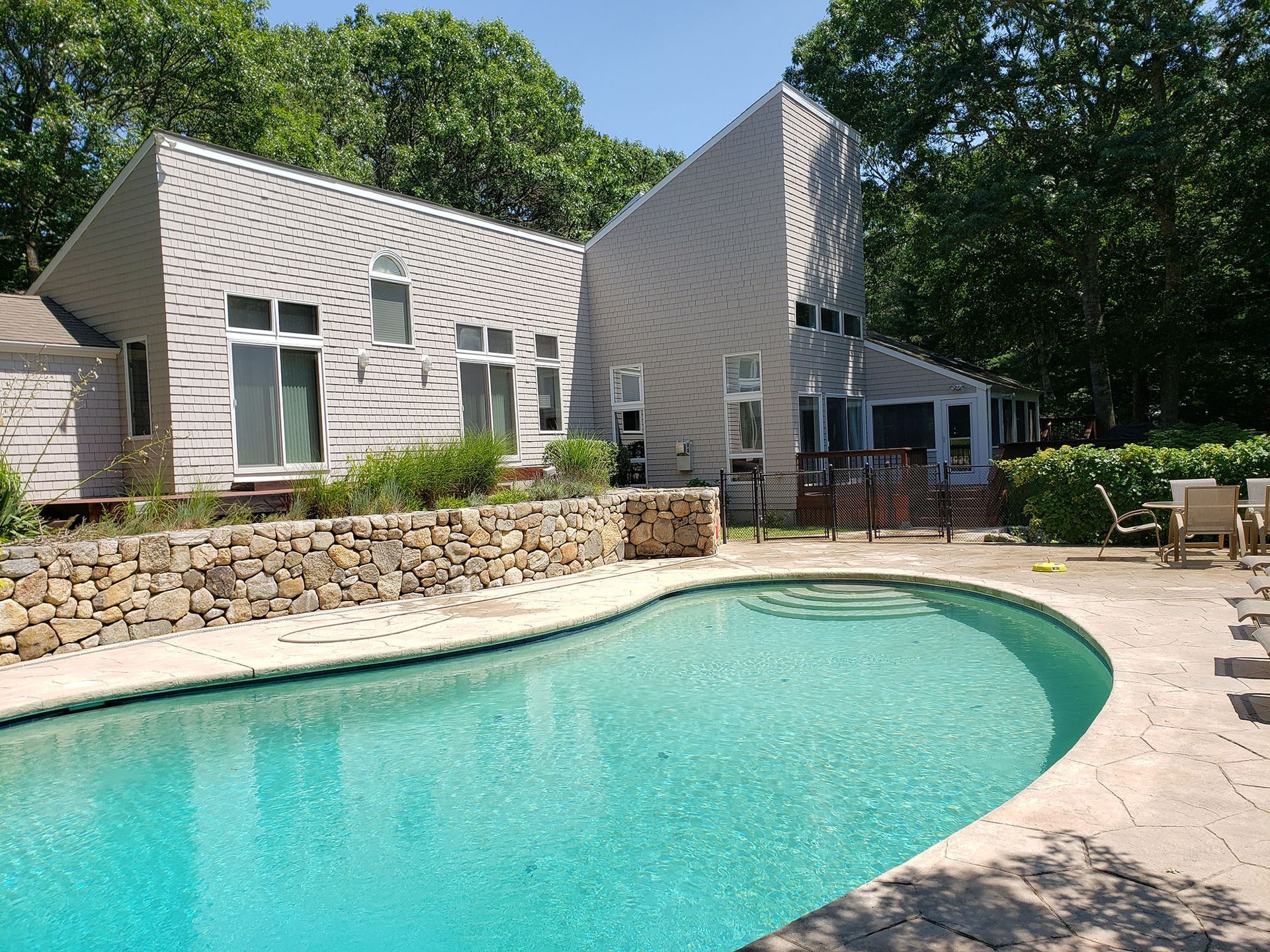Backyard swimming pool beside a stone patio and light-gray house under trees
